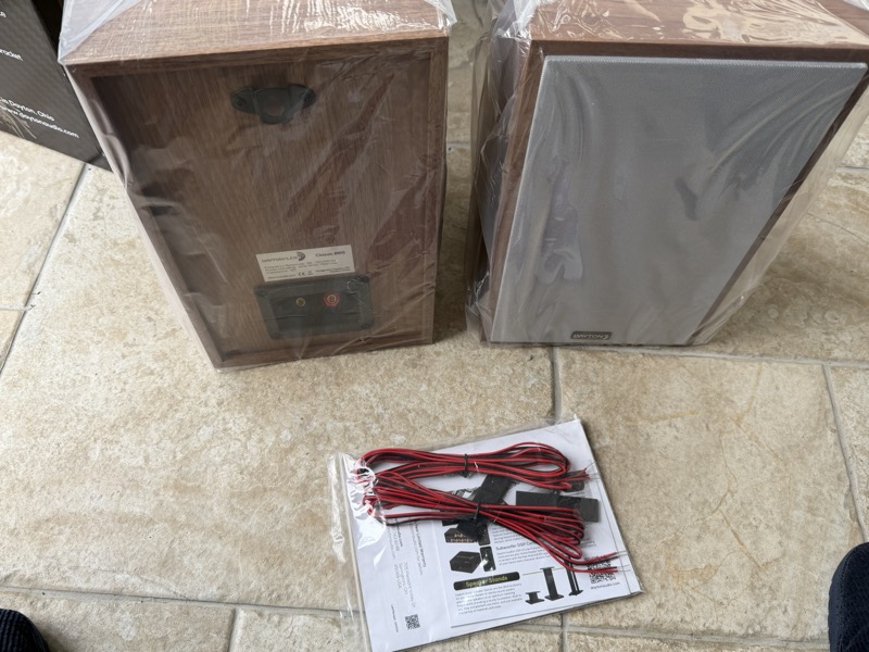 the speaker pair laid flat alongside a manual and a shorter set of speaker cables