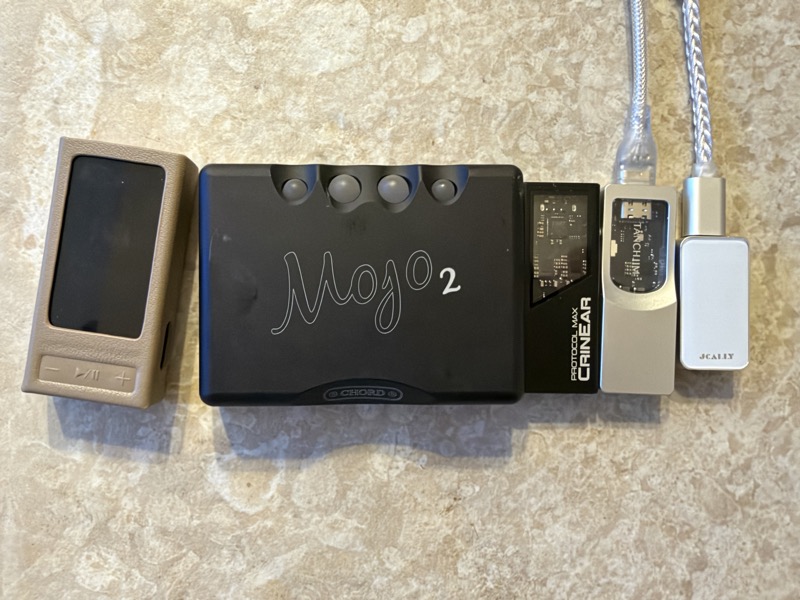 left to right: the fiio qx13, chord mojo 2, crinear protocol max, tanchjim space pro and a jcally dongle on a stone surface
