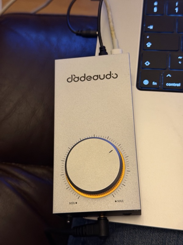 the Gigas1 top panel beside a MacBook, showing the large knurled volume knob and its orange backlight ring